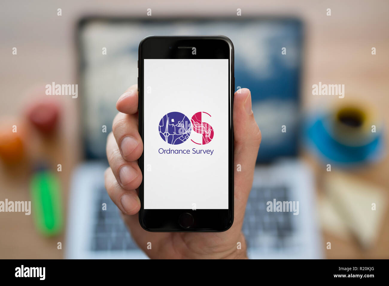 A man looks at his iPhone which displays the Ordnance Survey (OS) logo, while sat at his computer desk (Editorial use only). Stock Photo