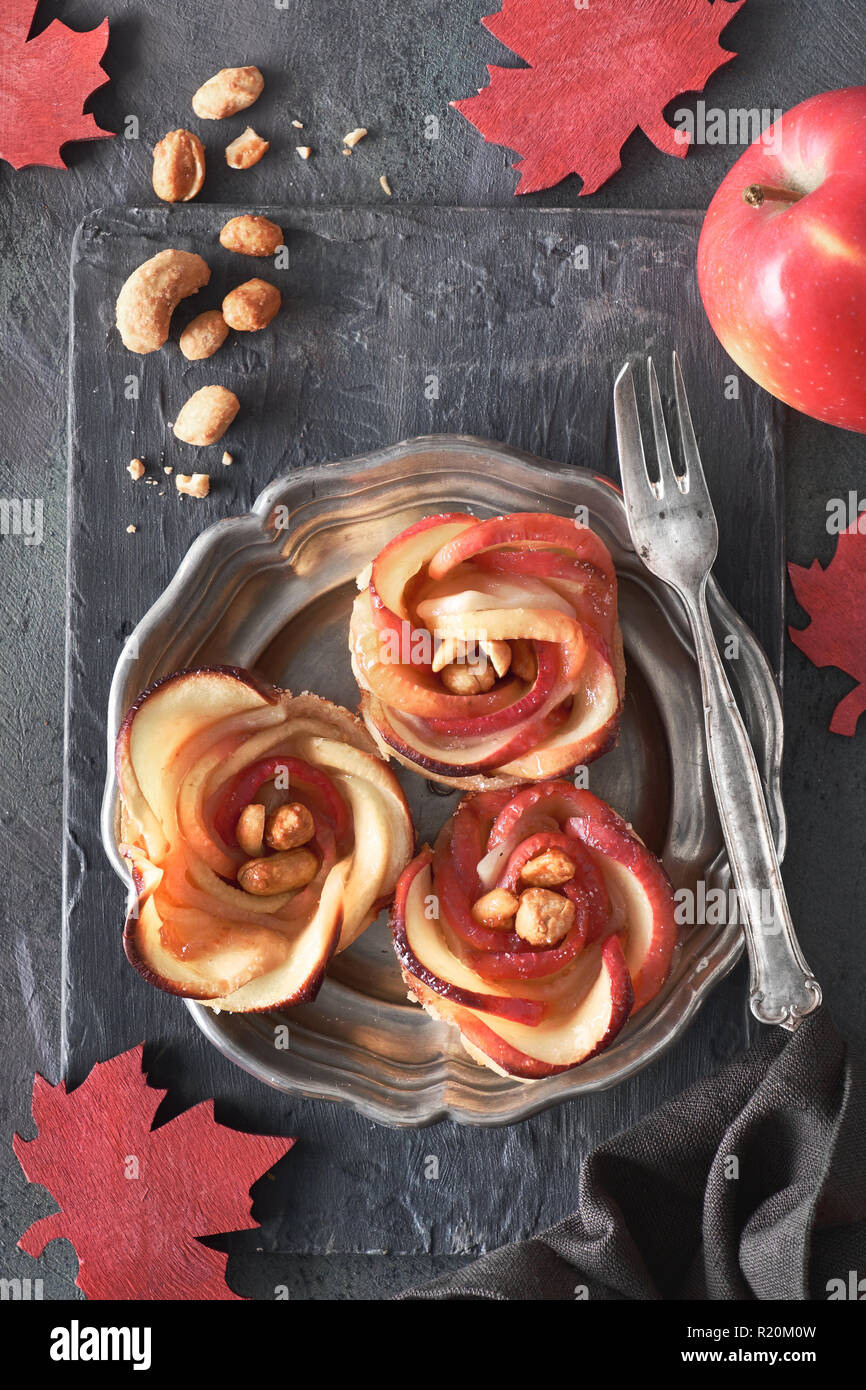 Three homemade puff pastries with rose shaped apple slices on metal plate. Top lay on wooden board with red maple leaves and caramelized nuts. Stock Photo