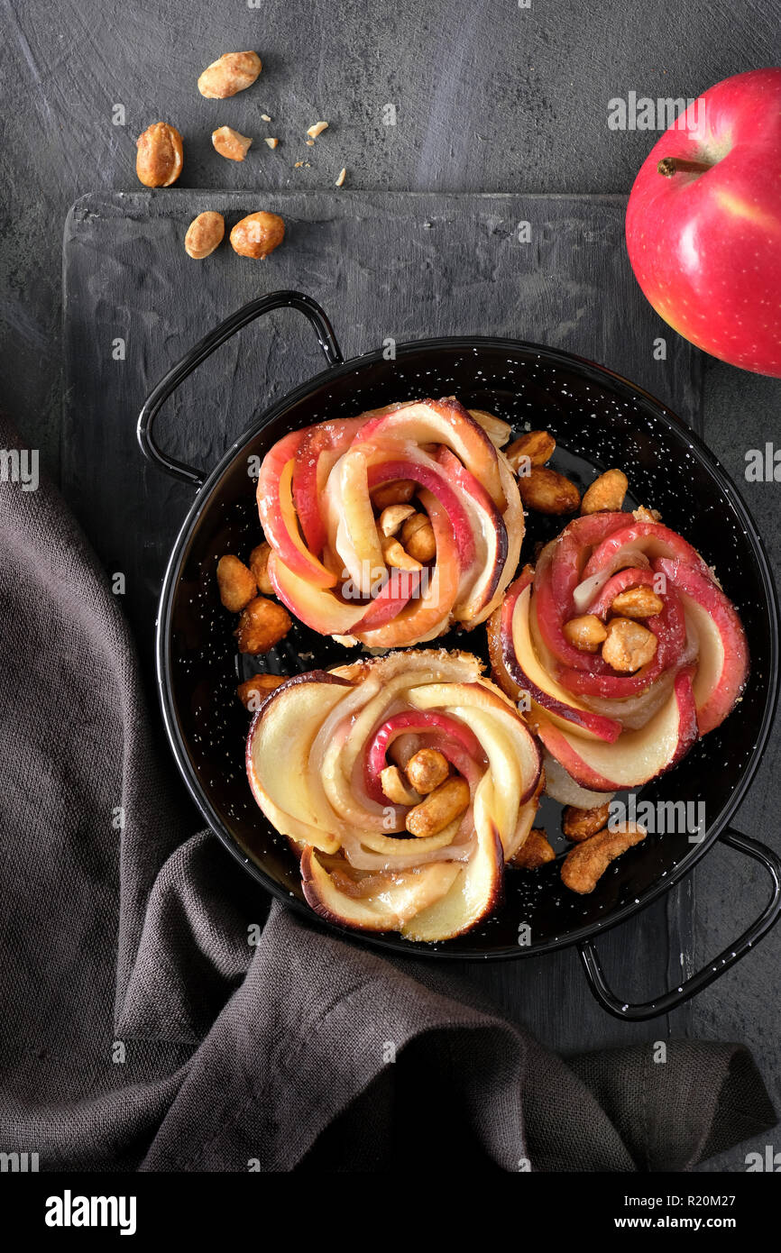 Three puff pastries with rose shaped apple slices baked in metal skillet and a fersh red apple. Top lay on dark background with linen towel and carame Stock Photo
