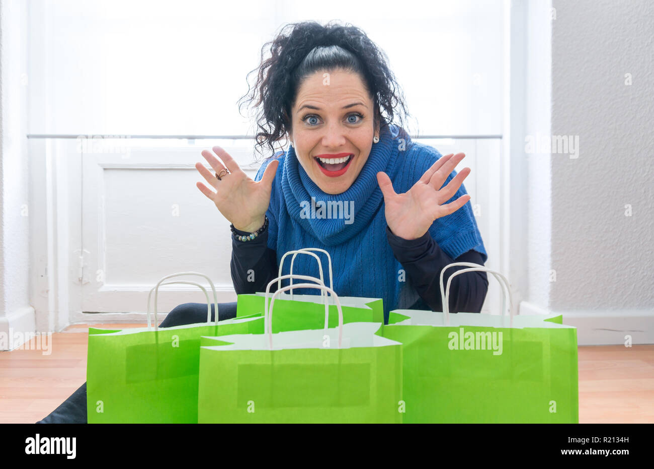 middle age woman with blue eyes white skin and black hair making a funny surprise expression looking at the camera. Many green shopping bags full of christmas gifts from winter sales Stock Photo