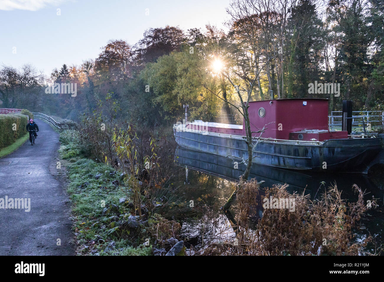 Sunlight bursts through the trees and mist on a cold frosty morning behind a canal barge with path alongside with cyclist. Lagan Towpath, Belfast, N.I Stock Photo