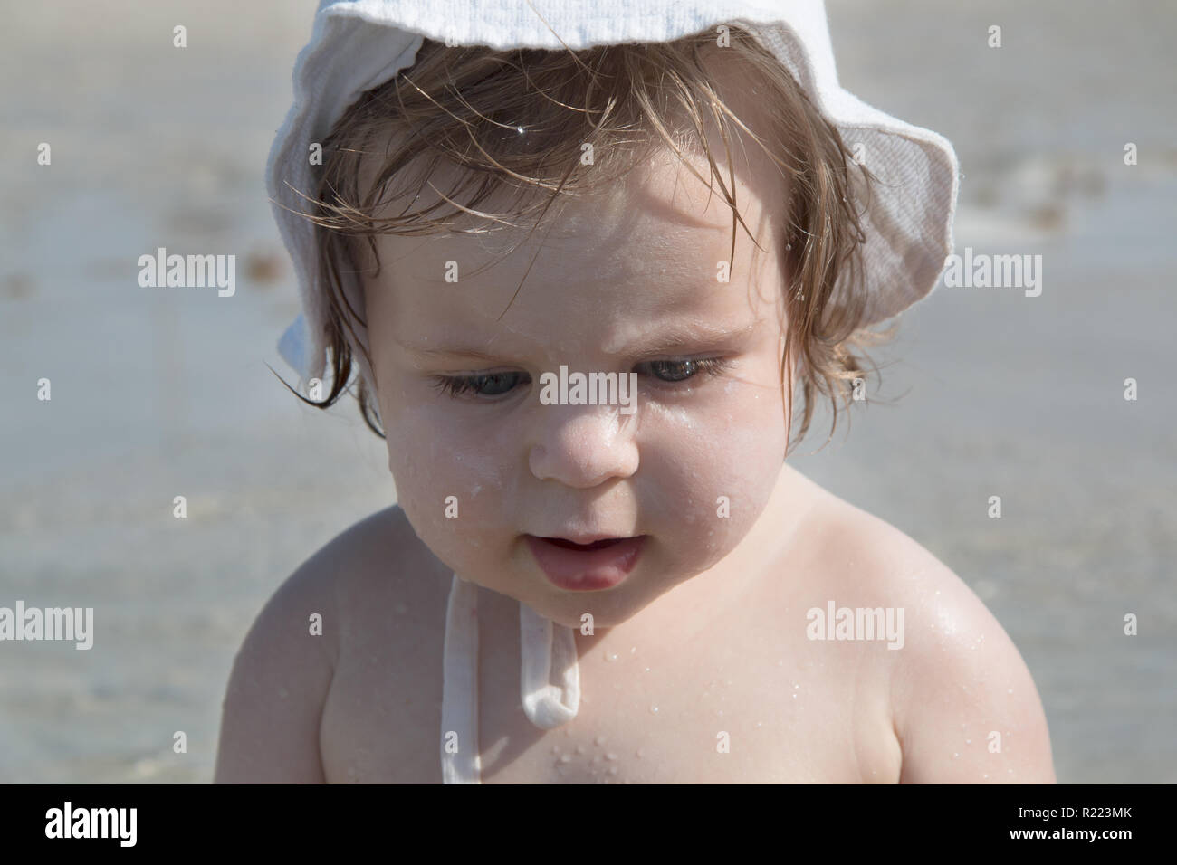 Cute sad baby with sunscreen cream on cheek against sea background. Pretty infant girl in white hat and with sunscreen and water drops on her face pla Stock Photo