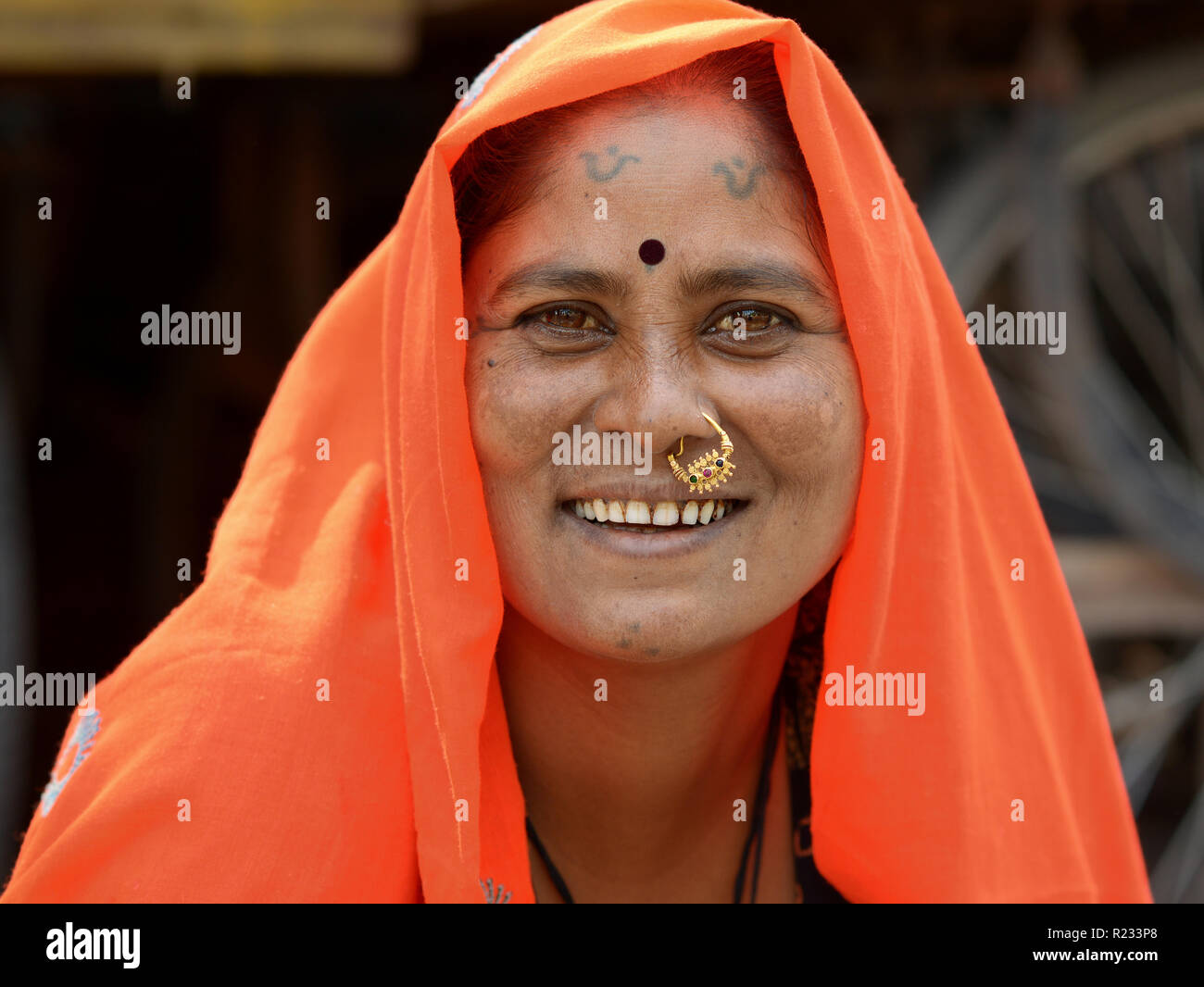 Middle-aged Rajasthani woman with traditional nose jewelry and tribal tattoos on chin and forehead wears an orange headscarf (dupatta). Stock Photo