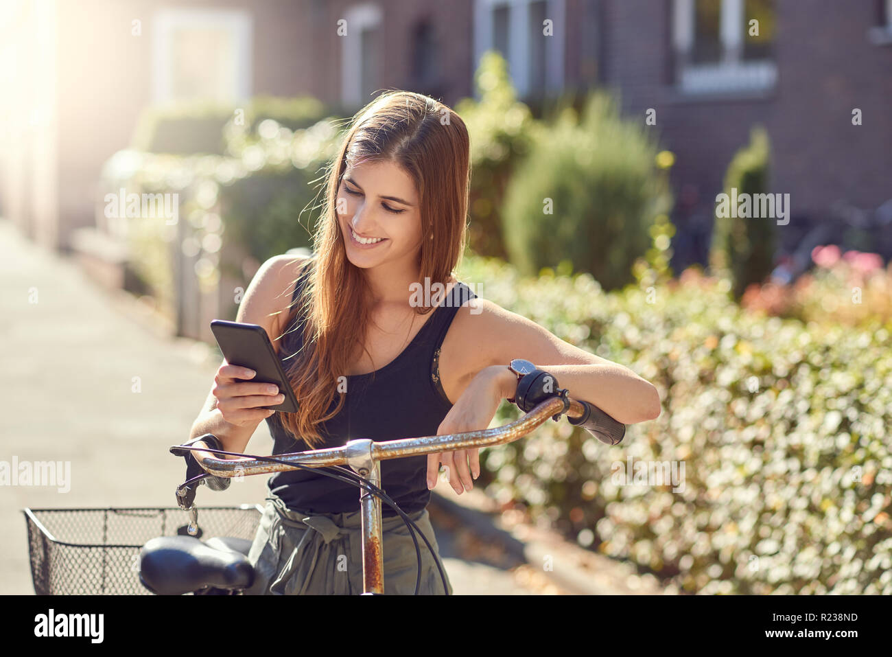 Beautiful young woman with long hair in half-length front portrait outdoors in the city standing leaning on vintage bicycle using modern smartphone, l Stock Photo
