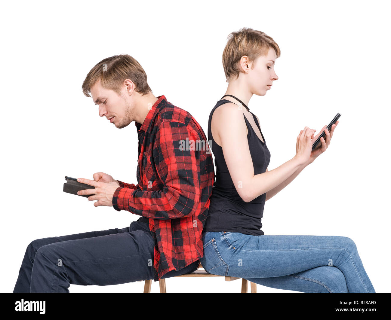 Young couple sitting on one chair back to back and looking at his smartphones. Isolated on white. Gadget addiction, millennials, hipsters, loneliness  Stock Photo