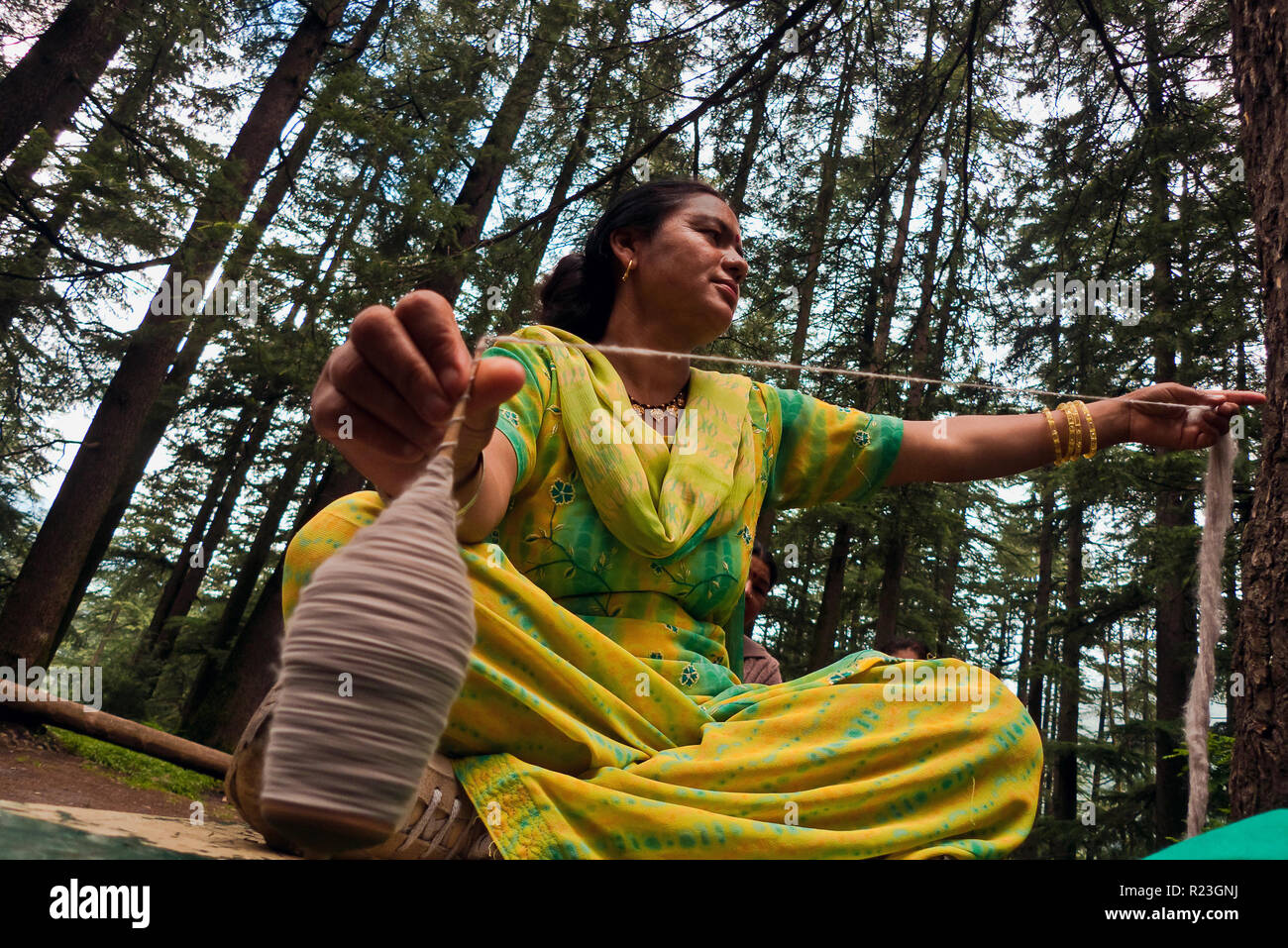 India, Himachal Pradesh, Manali: Indian woman weaves a ball of yarn sitting on a bench at the foot of a tree Stock Photo