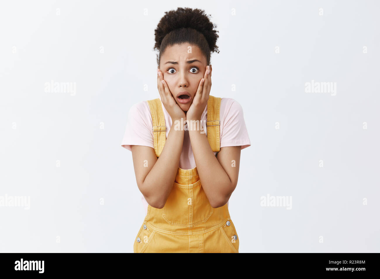 Woman hearing terrible disaster happened to friend. Portrait of shook intense worried female with dark skin in yellow overalls, gasping, losing speech Stock Photo