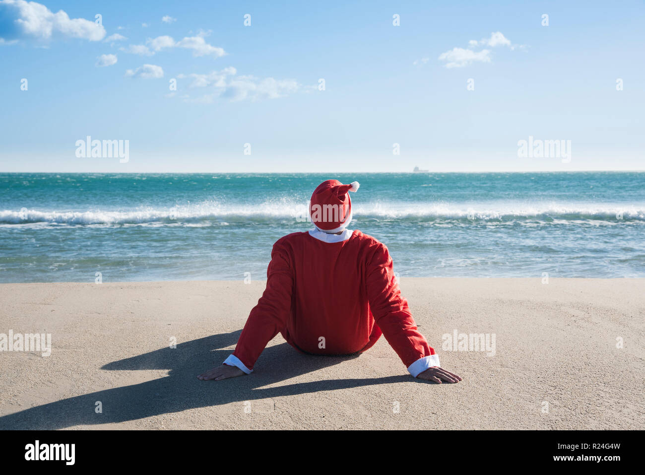 santa claus, seen from behind, sitting on the sand of a beach facing ...