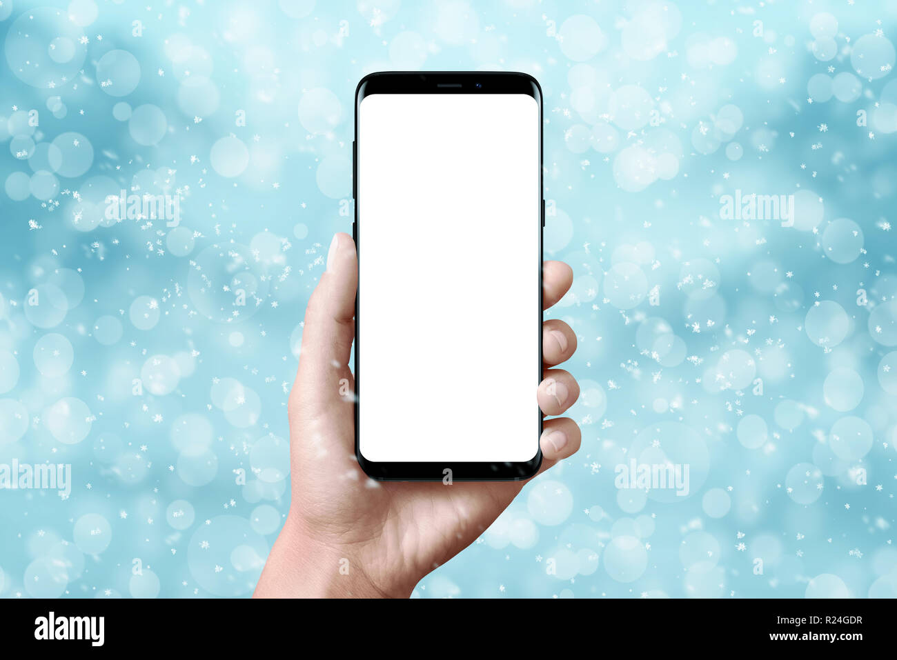 Modern black phone in hand. Isolated, blank screen for mockup. Christmas scene with blue background with snowflakes, light and bokeh. Stock Photo