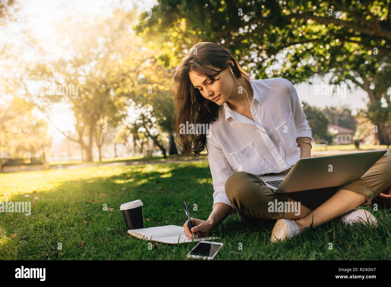businesswoman sitting on grass at park with laptop and making notes in her diary. Freelancer female working while sitting at park. Stock Photo