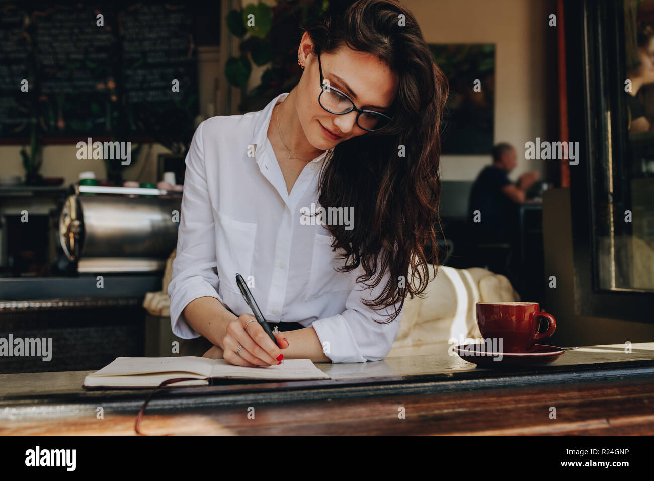 Beautiful woman sitting near a window at cafe and making some important notes in her book. Caucasian female writing in book at coffee shop. Stock Photo