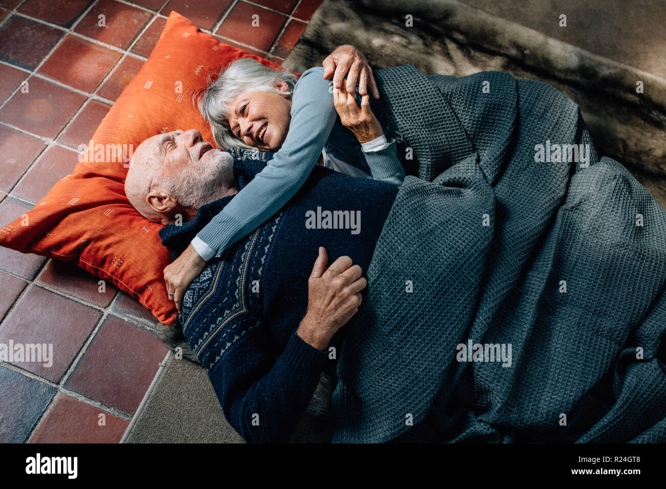 Senior couple lying down on floor holding each other and talking. Old man and woman sleeping on floor at home covering a blanket and talking. Stock Photo