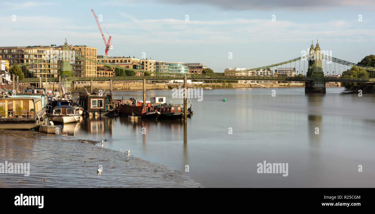 London, England, UK - September 9, 2018: Houseboats are moored at a jetty on the River Thames beside the Hammersmith Suspension Bridge in West London. Stock Photo