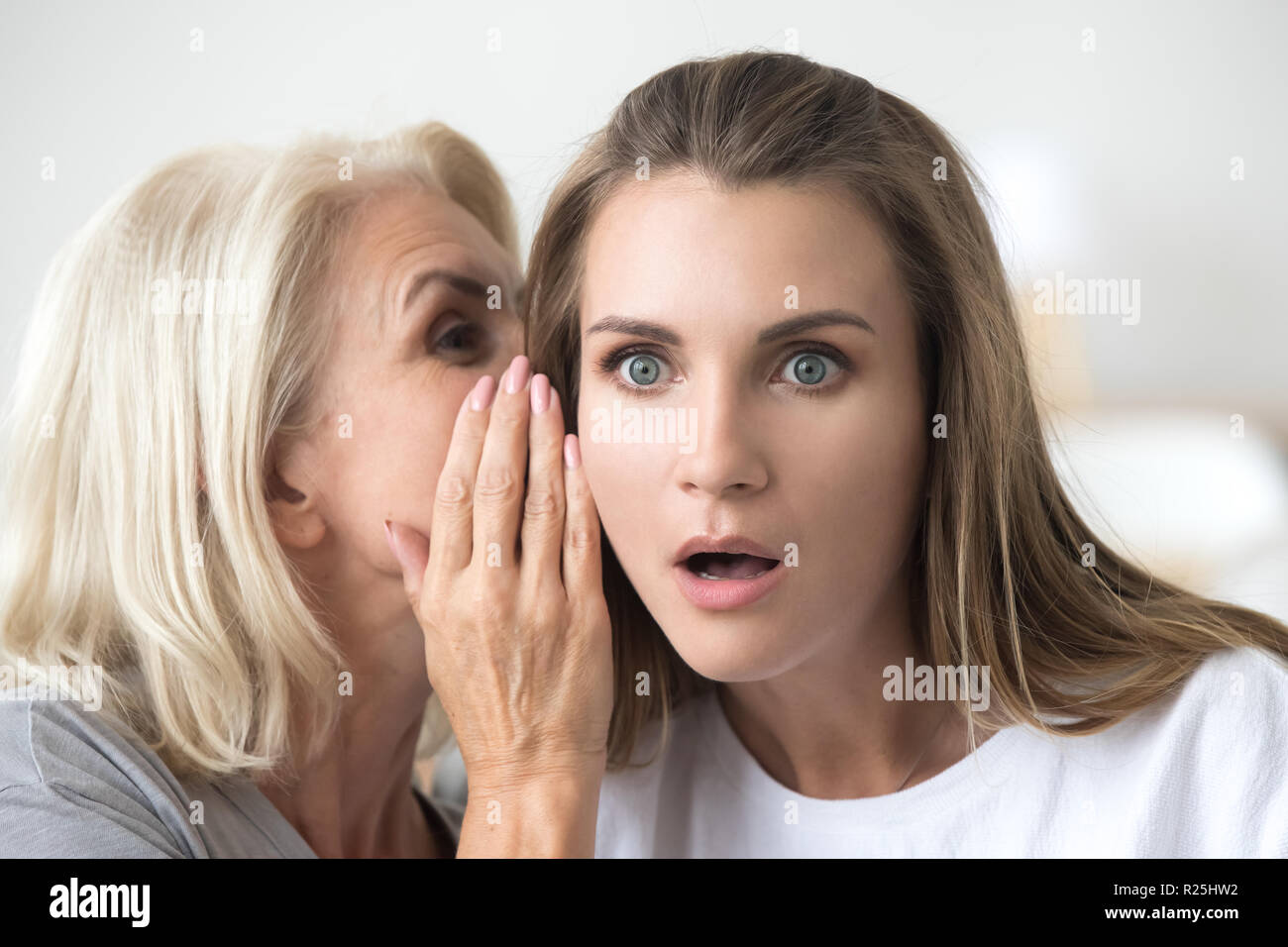 Older mother whispering in young daughter ear telling shocking n Stock Photo