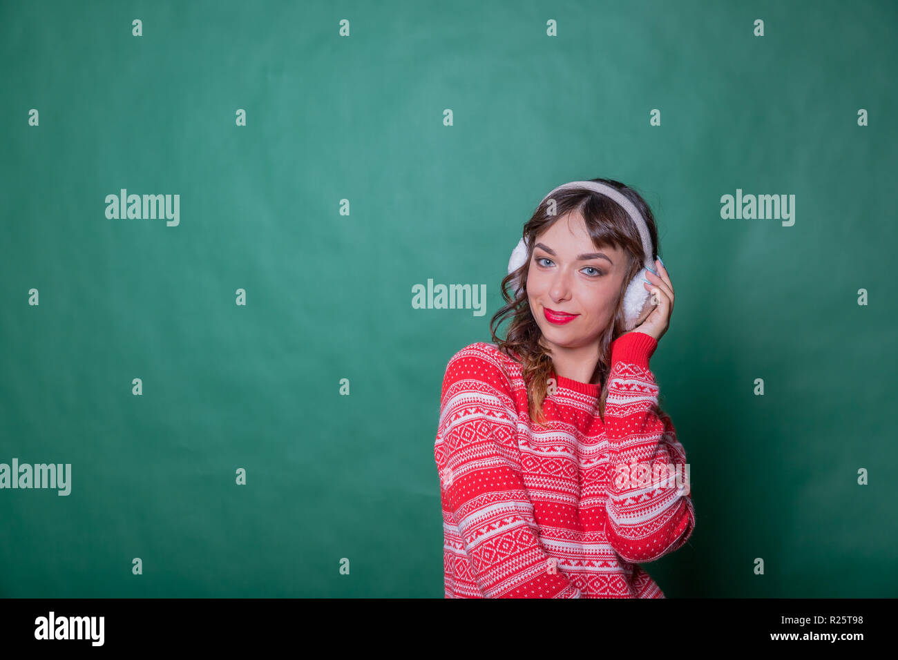 Winter Beauty Woman on green wall background. Winter holidays concept. Skin and hair care in cold season. Beautiful woman with long hair wearing sweater and earmuffs.Copy space Stock Photo