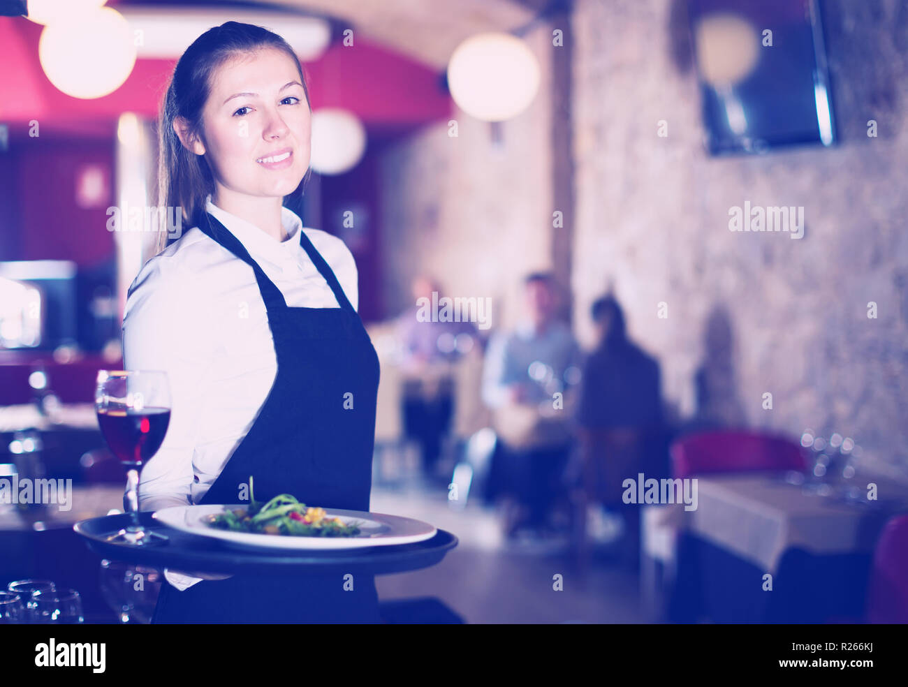 Portrait of polite waitress wearing apron holding tray with dishes in ...