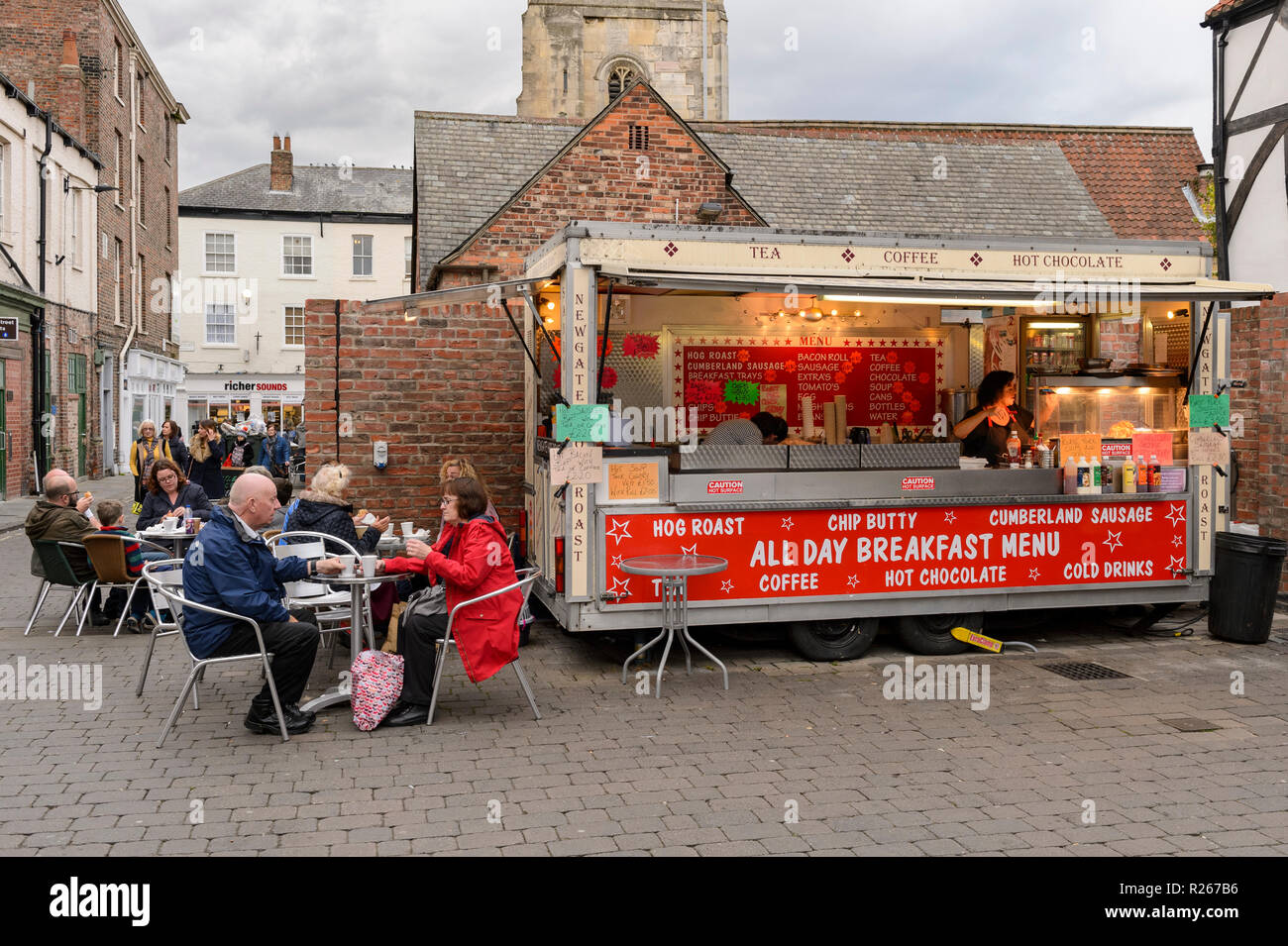 Customers sit outside at tables, eating street food & drinking & 2 women work in Newgate Hog Roast van - Shambles Market, York, Yorkshire, England, UK Stock Photo