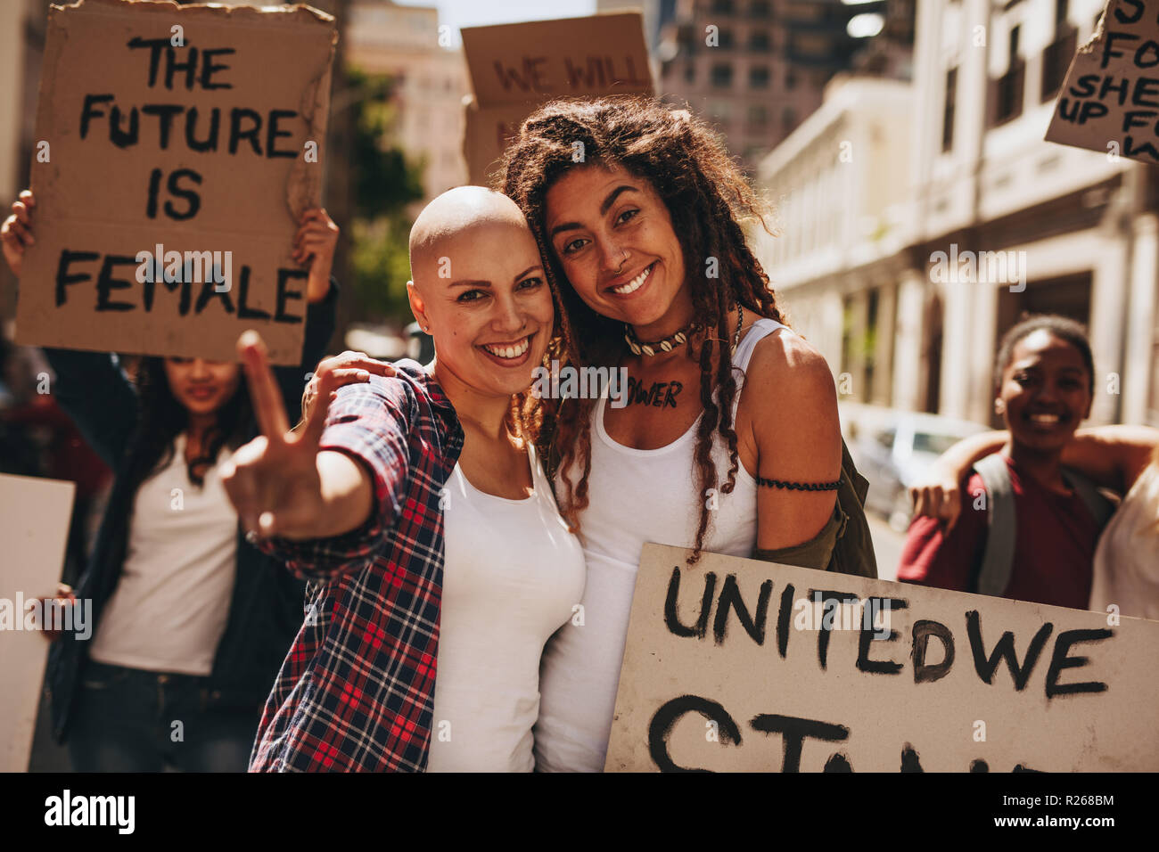 Happy female activists protesting outdoors with placards showing peace sign. Group of demonstrators enjoying outdoors in road with protest. Stock Photo