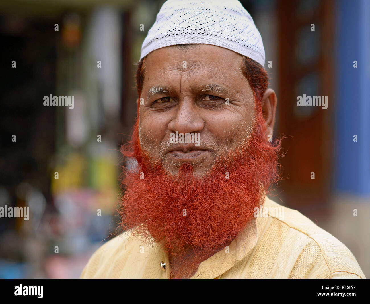 Elderly Indian Muslim man with henna-dyed Islamic beard wears a white prayer cap (taqiyah) and smiles for the camera. Stock Photo