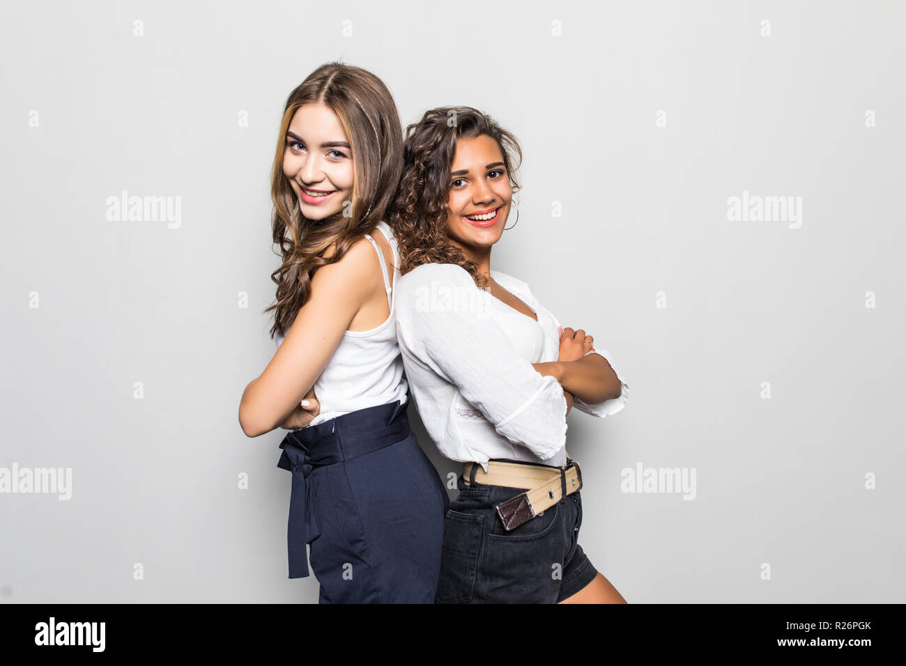 Two young girls stand back to back and smiling to camera on gray ...