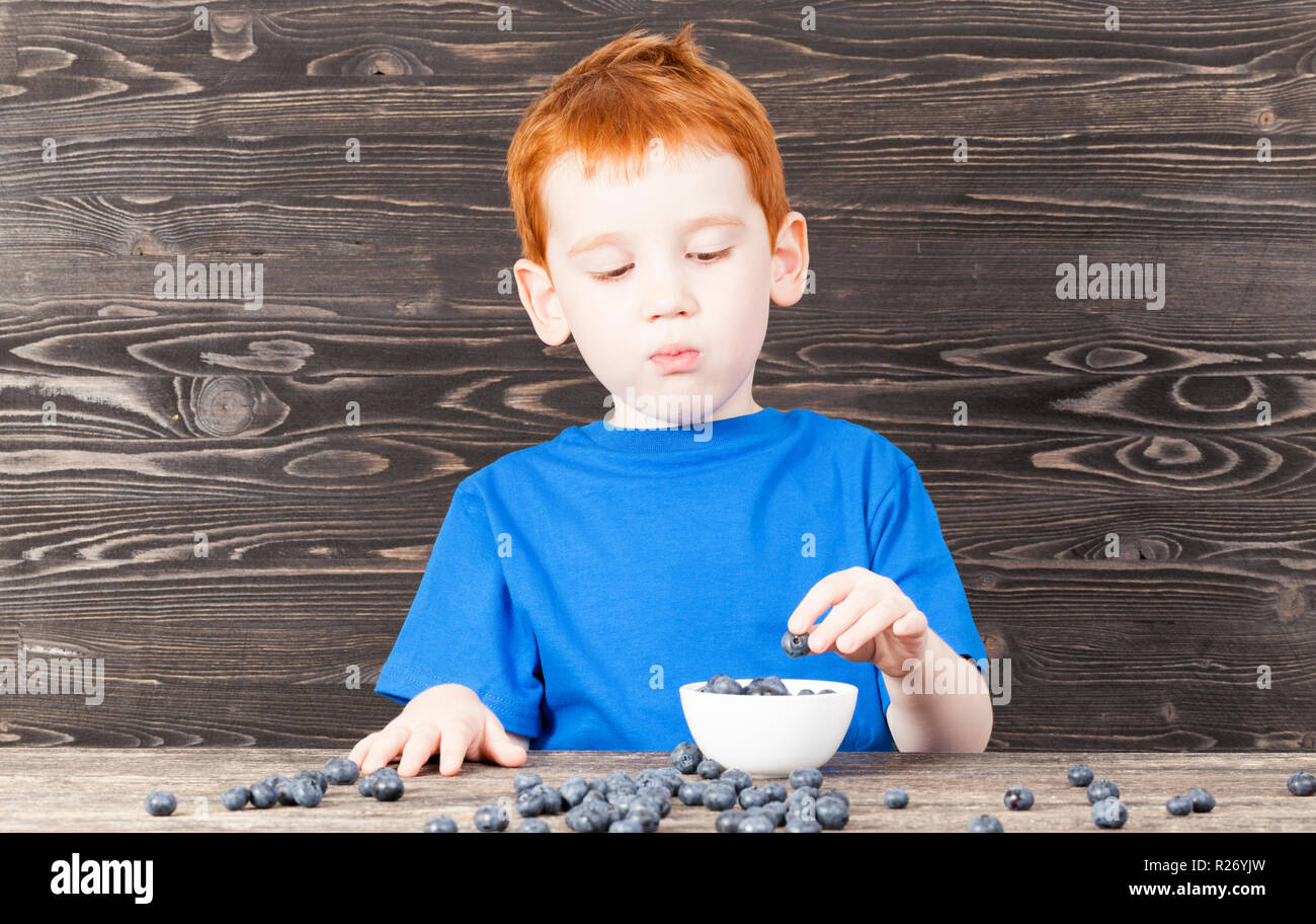 a boy with red hair and freckles puts blueberry berries in a plate, in the kitchen, a black kitchen table Stock Photo