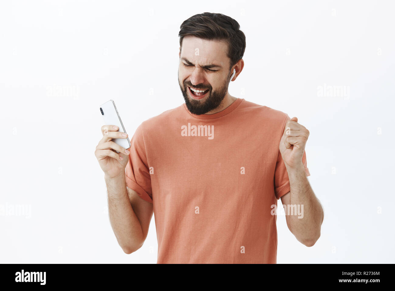 Waist-up shot of enthusiastic pleased and delighted carefree charming adult male in wireless earphones holding smartphone dancing in rhythm of music s Stock Photo