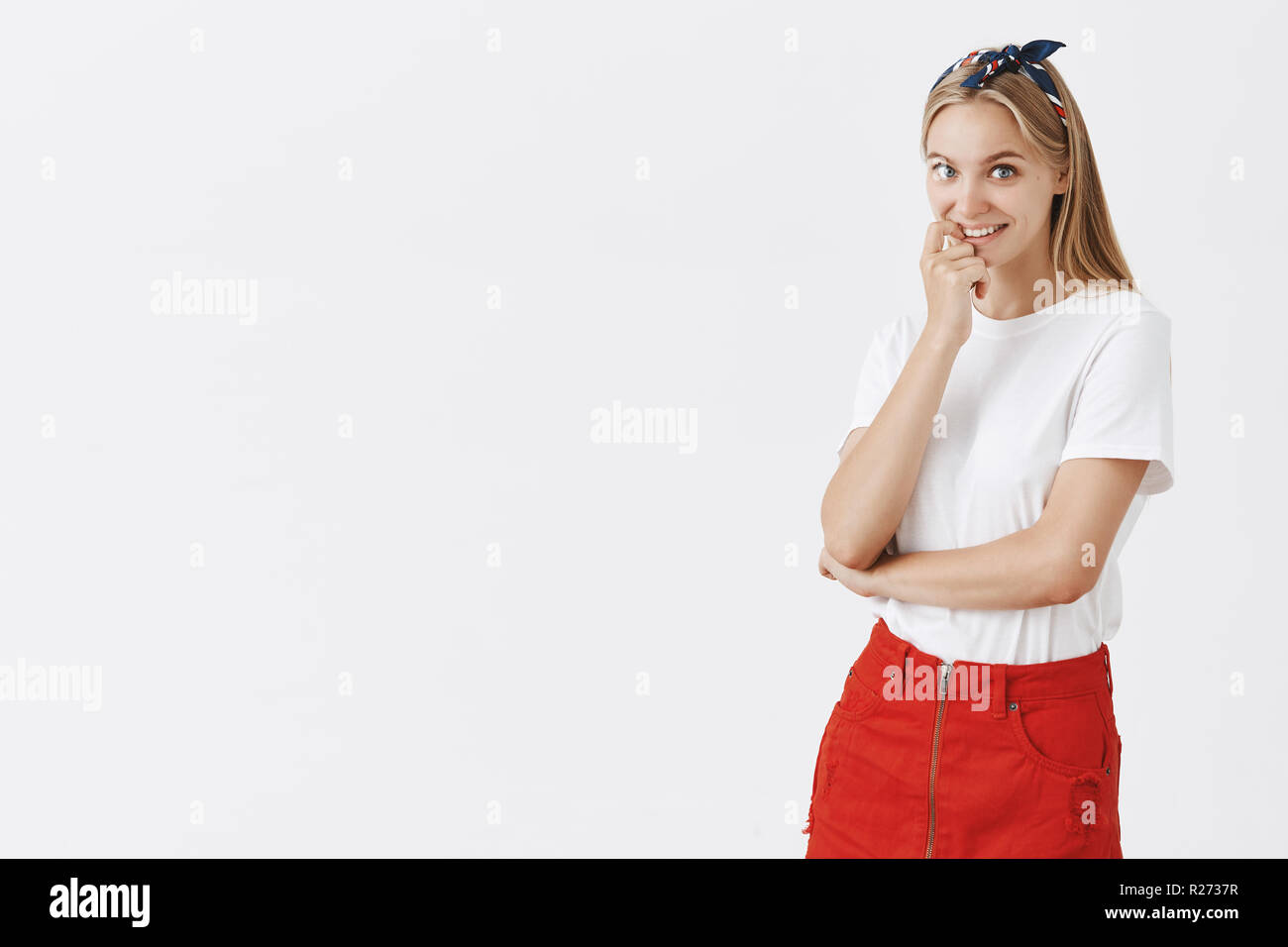 Studio shot of curious good-looking stylish caucasian girl with fair hair in headband and trendy red skirt, biting fingernail and smiling wanting some Stock Photo