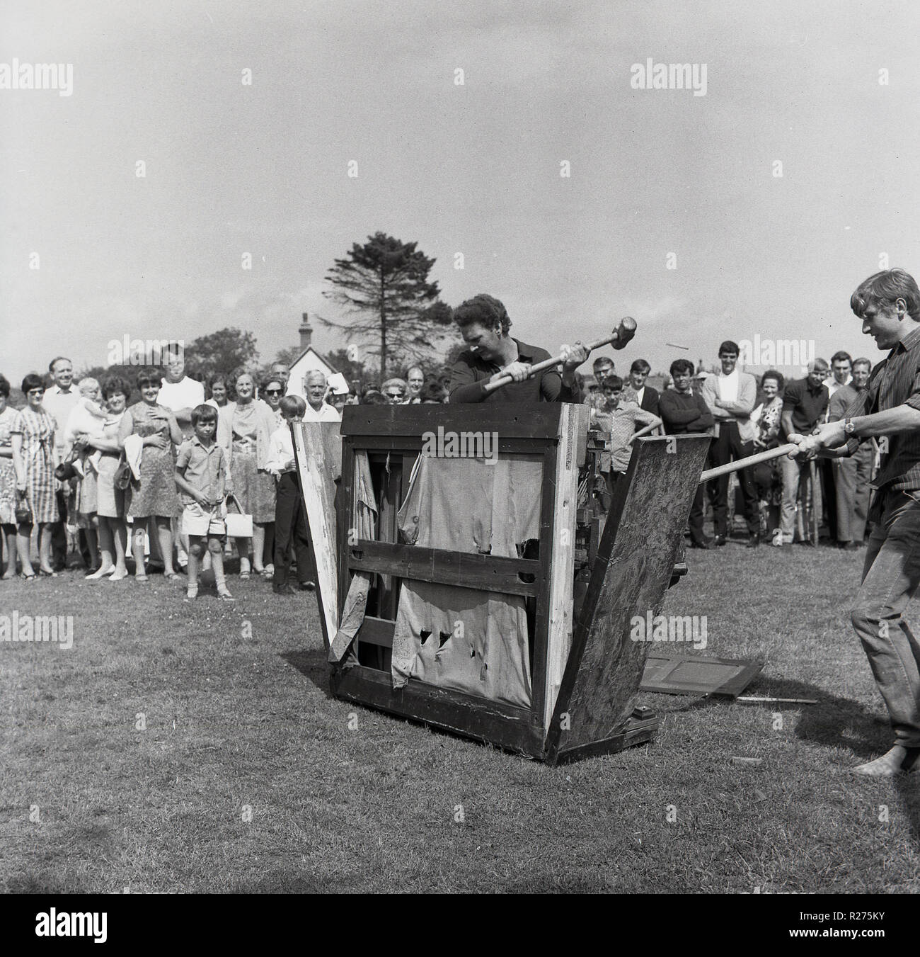 1965, historical, men smashing up an upright piano outside in a field at a village fete, Oxfordshire, England, UK. Piano smashing contests were a popular features of British village fetes in this era, with teams using sledgehammers to destroy the pianos into small enough peices that could be passed through a 9-inch diameter hole, with the winners doing it in the fastest time. Stock Photo