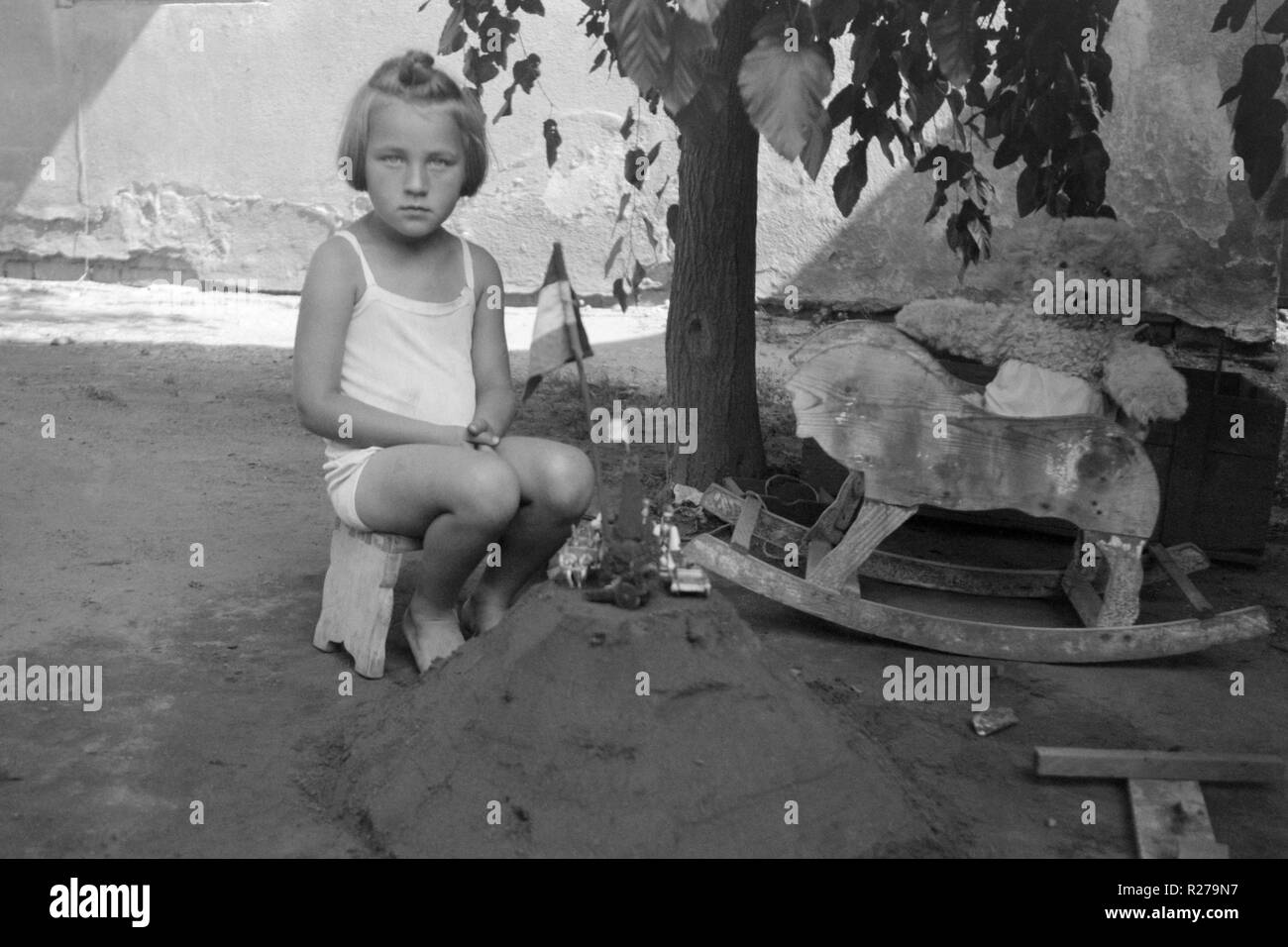 young girl sat on a stool in the garden next to a sandcastle and rocking horse 1930s hungary Stock Photo