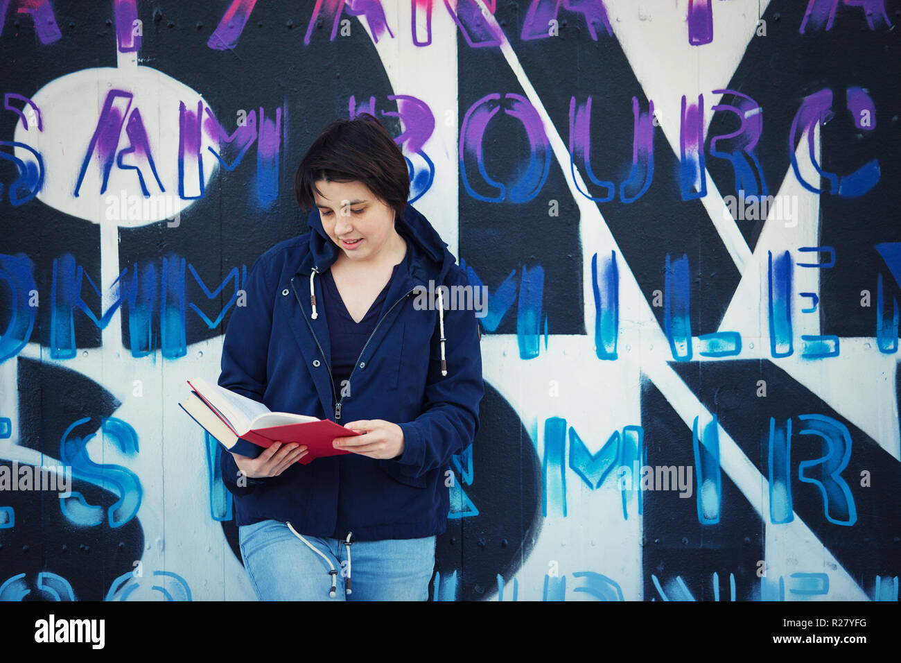 Casual woman student reading book, outdoors lifestyle portrait leaning back on graffiti wall background. Stock Photo
