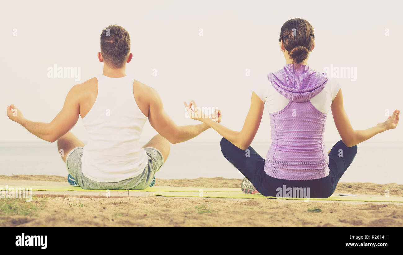 Positive cross-legged guy and girl training yoga poses on beach Positive cross-legged guy and girl training yoga poses on beach