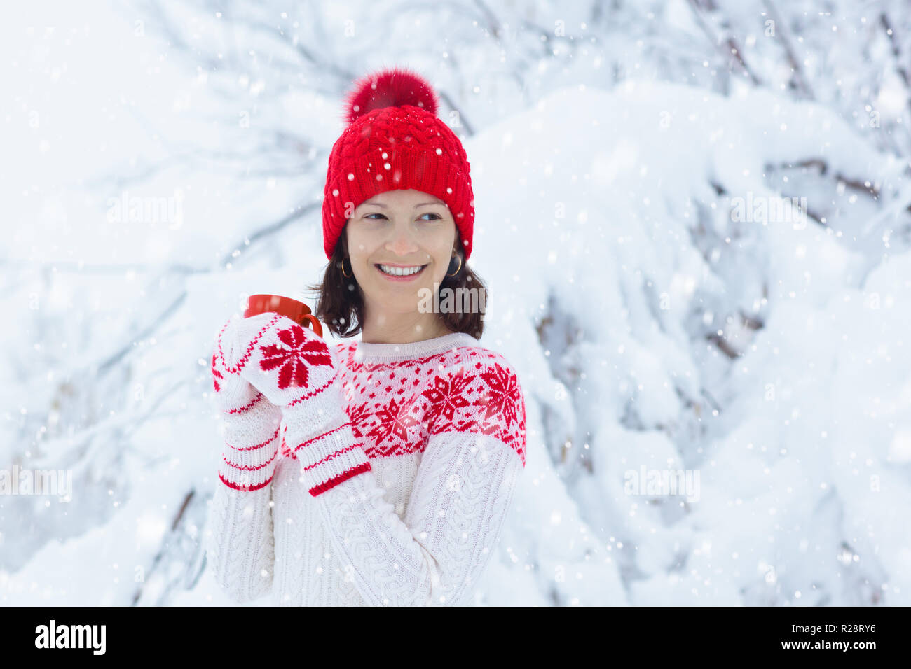 Woman drinking hot chocolate in Christmas morning in snowy garden. Girl in knitted Nordic sweater, hat and mittens holding cup with cocoa. Snow and wi Stock Photo