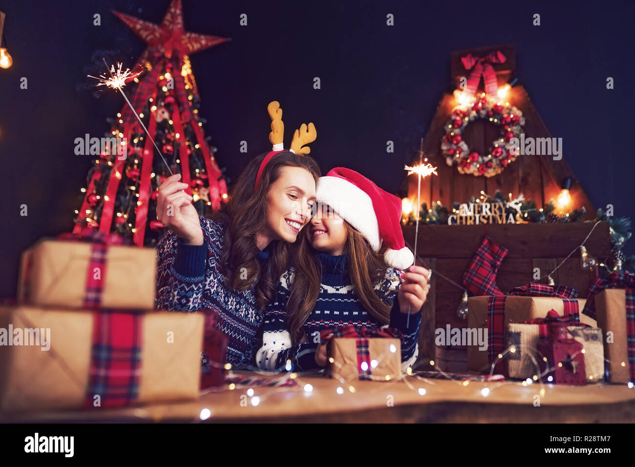 Happy mother in sweater, Christmas deer antlers and little girl in santa helper hat with sparklers in hands, gift boxes on foreground. Having fun over living room, christmas tree, fireplace background Stock Photo