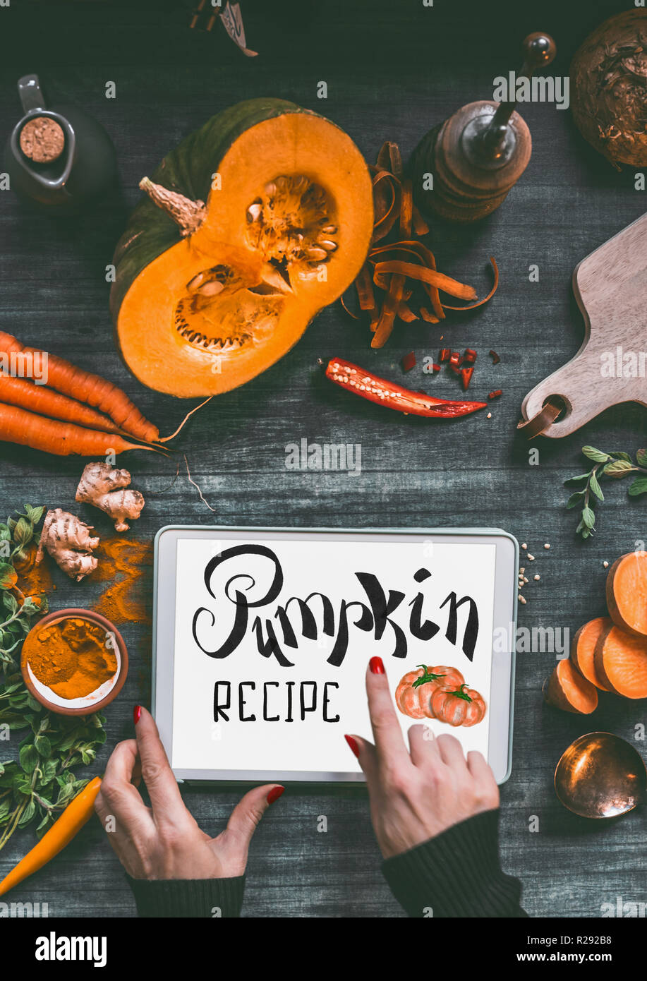 Female hand pointing with finger on tablet pc with text lettering: Pumpkin recipe on kitchen table background with orange cooking ingredients: sweet p Stock Photo