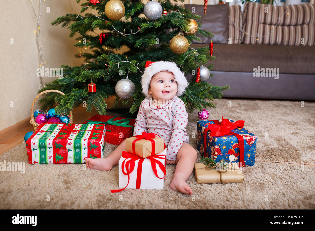 Happy baby girl in Santa Claus hat holding Christmas gifts at christmas tree at home Stock Photo