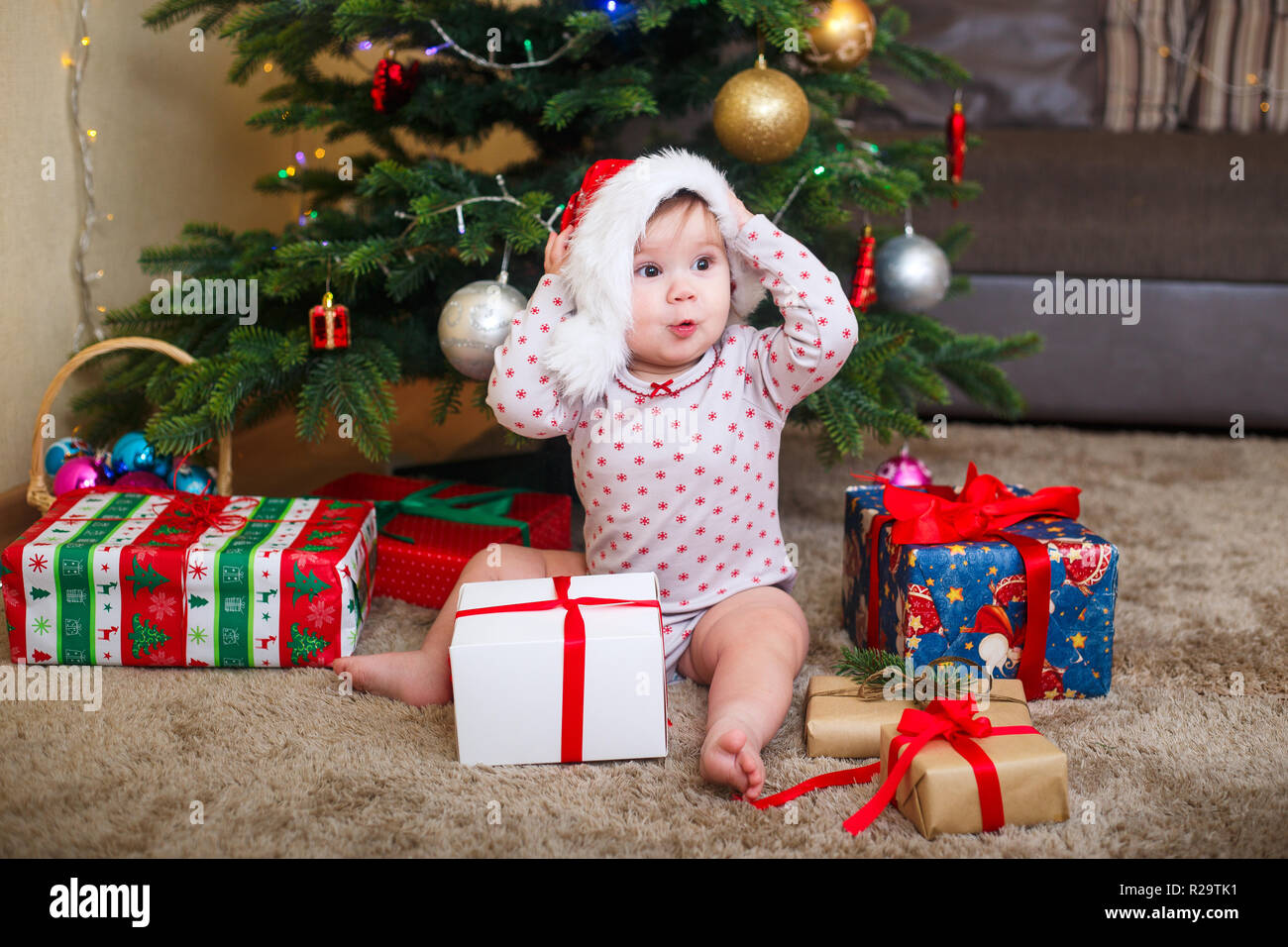 Happy cute baby girl in Santa Claus hat holding Christmas gifts at christmas tree at home Stock Photo