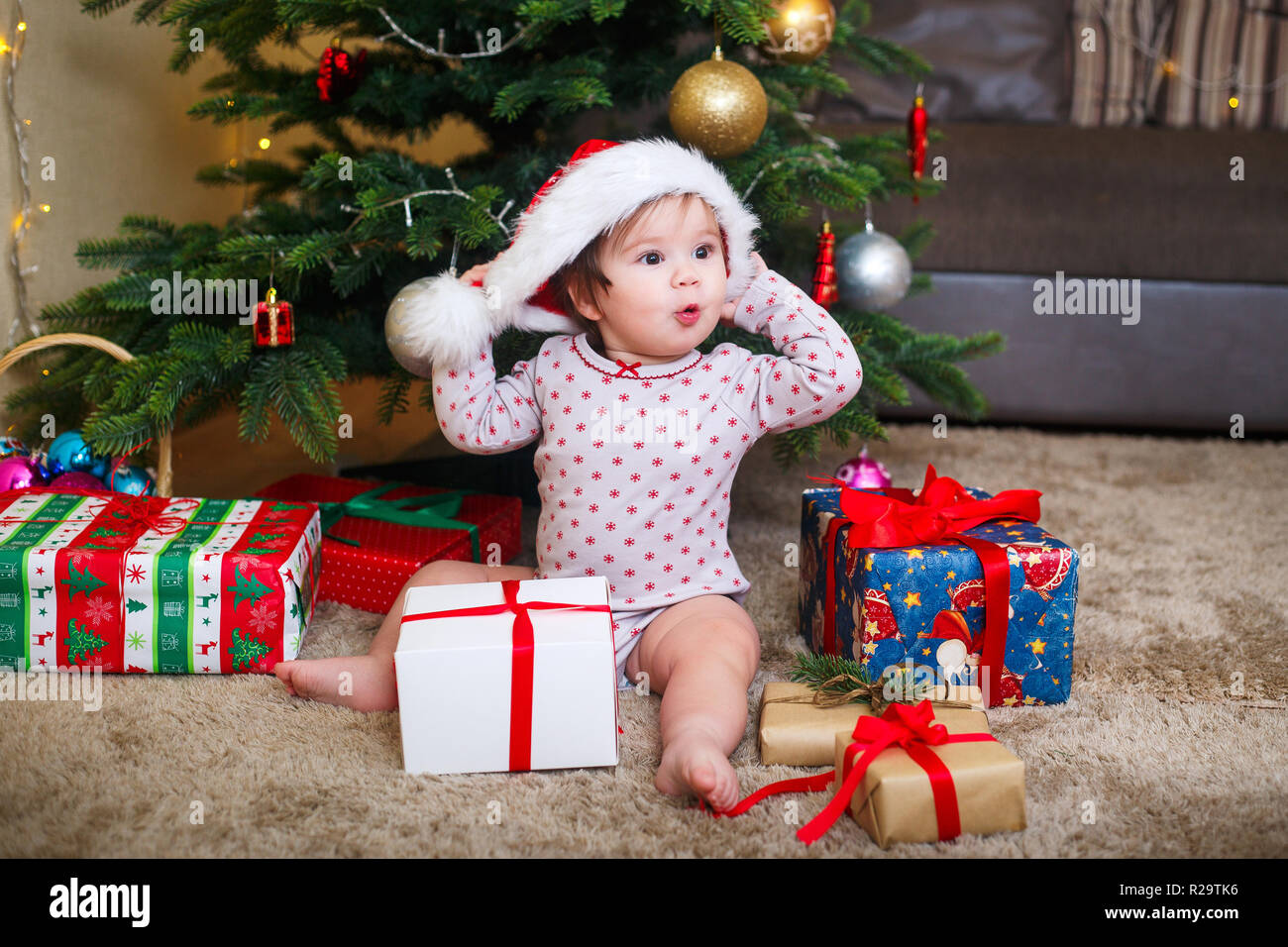 Happy cute baby girl in Santa Claus hat holding Christmas gifts at christmas tree at home Stock Photo