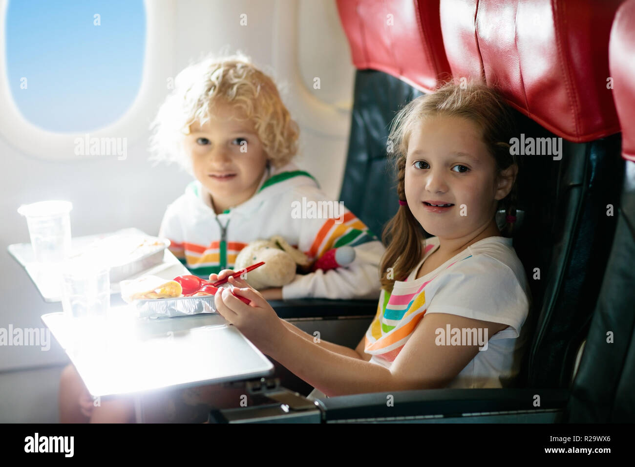 Child in airplane window seat. Kids flight meal. Children fly. Special inflight menu, food and drink for baby and kid. Girl and boy eating healthy lun Stock Photo