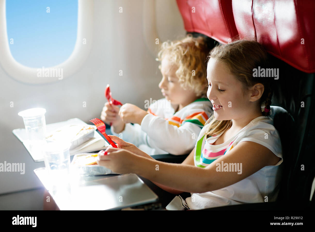 Child in airplane window seat. Kids flight meal. Children fly. Special inflight menu, food and drink for baby and kid. Girl and boy eating healthy lun Stock Photo