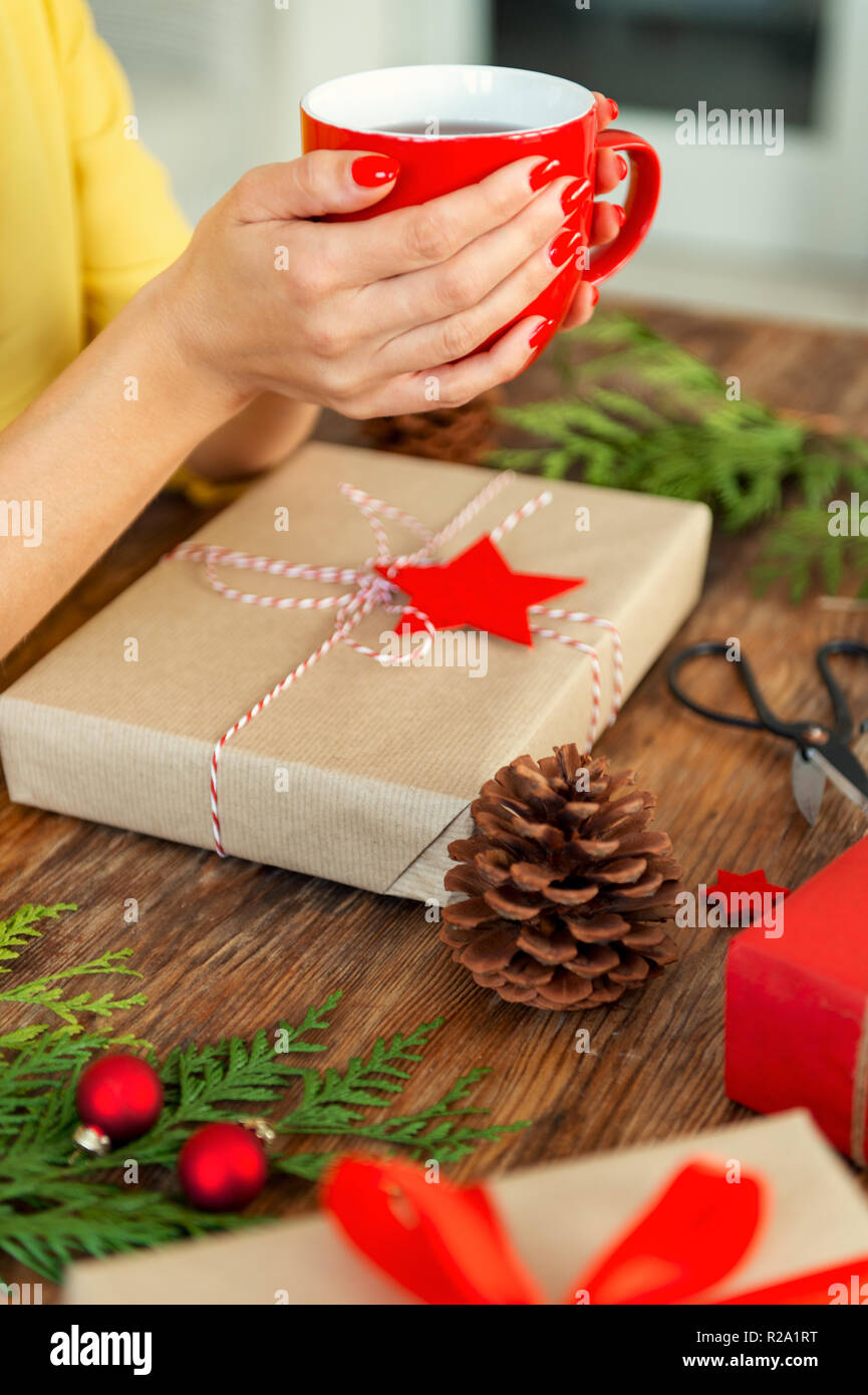 Unrecognisable woman sitting at a table with beautifully wrapped christmas present, holding cup of tea. Gift wrapping xmas concept. Stock Photo