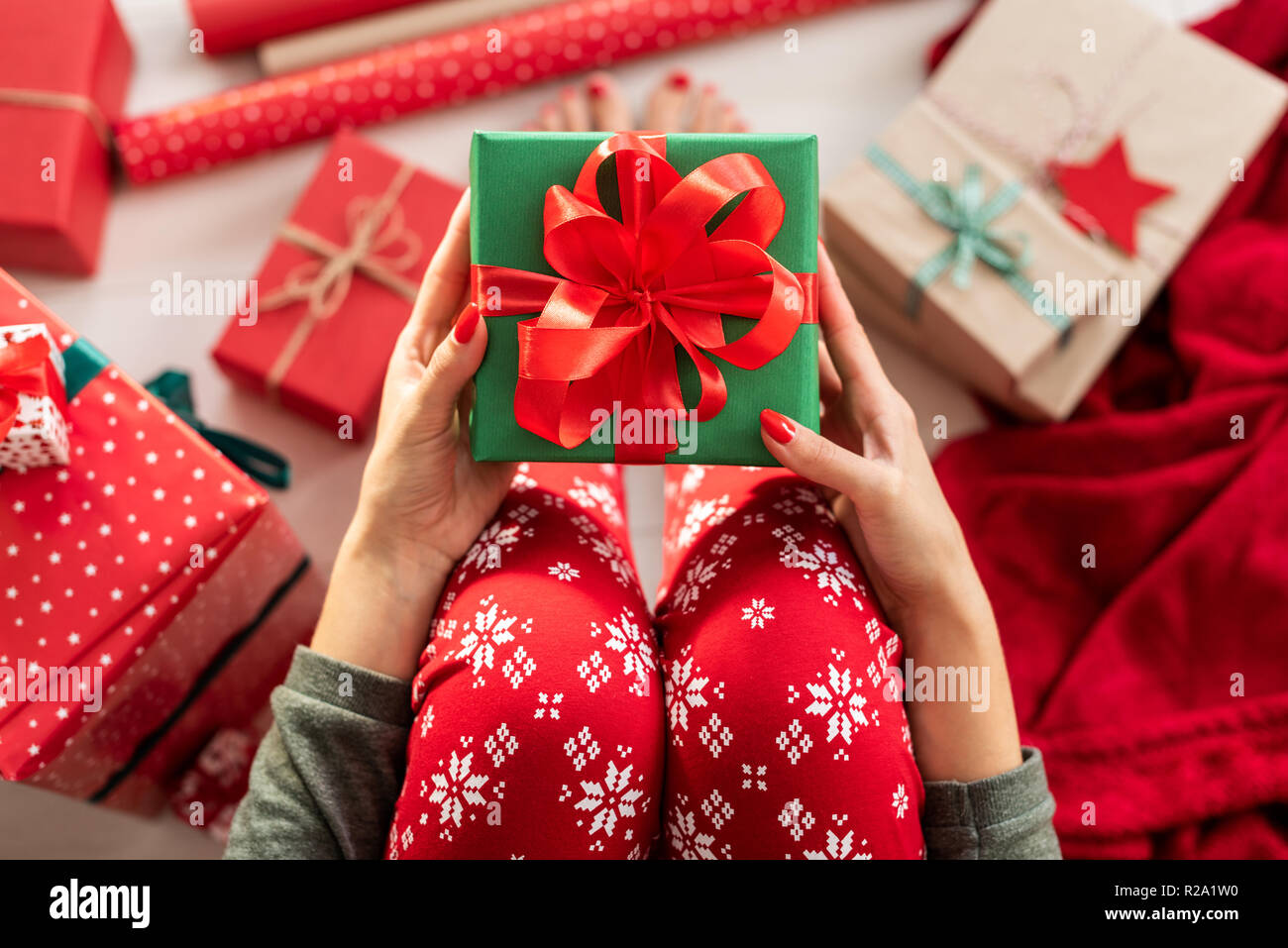 Young woman wearing xmas pajamas sitting on the floor amongst wrapped christmas presents, holding beautiful gift box with large red ribbon bow. Person Stock Photo
