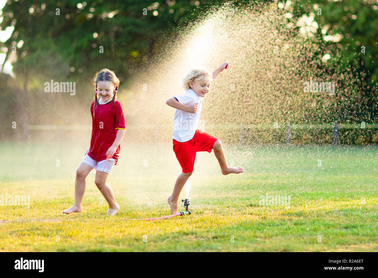 Kids play with water on hot summer day. Children with garden sprinkler. Outdoor fun. Boy and girl run on football field after training under water dro Stock Photo