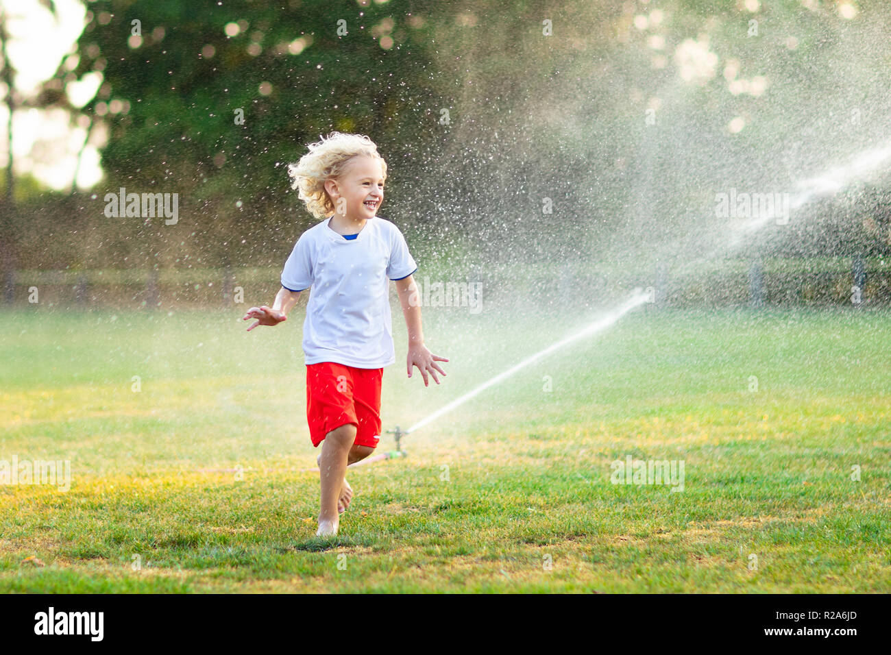 Kids play with water on hot summer day. Children with garden sprinkler. Outdoor fun. Boy and girl run on football field after training under water dro Stock Photo