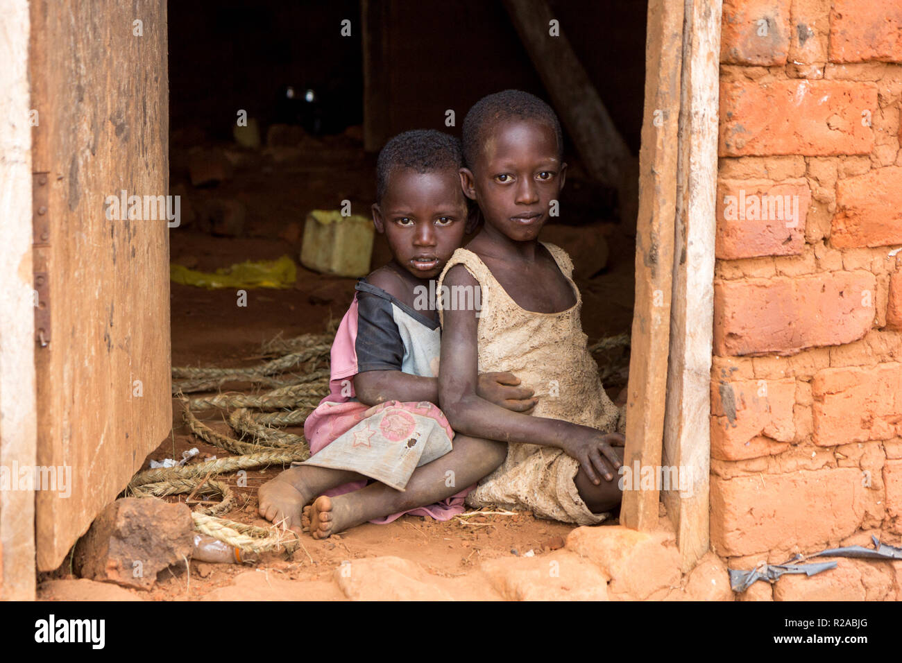 Two Ugandan children in ragged clothes sitting in an embrace on a dirt floor at the doorstep of a house. Stock Photo