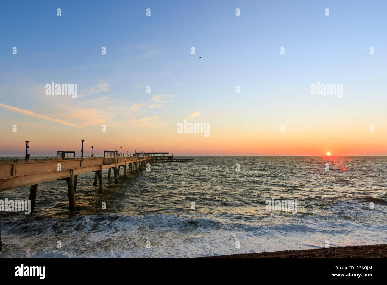 Sunrise over the sea and Deal Pier in Kent, England. High tide with rough sea and waves crashing onto the shingle beach. Thin band of orange sky on horizon with blue sky above. Stock Photo