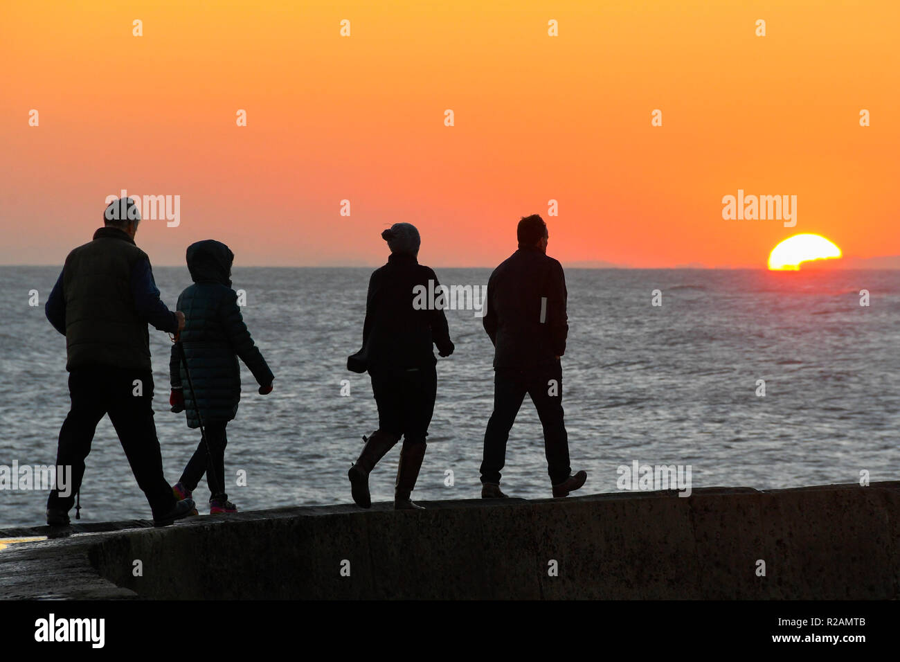 Lyme Regis, Dorset, UK.  18th November 2018. UK Weather. People walking along the Cobb Harbour wall silhouetted against the sunset  at the end of a clear sunny autumn day at the seaside resort of Lyme Regis in Dorset.  Picture Credit: Graham Hunt/Alamy Live News Stock Photo