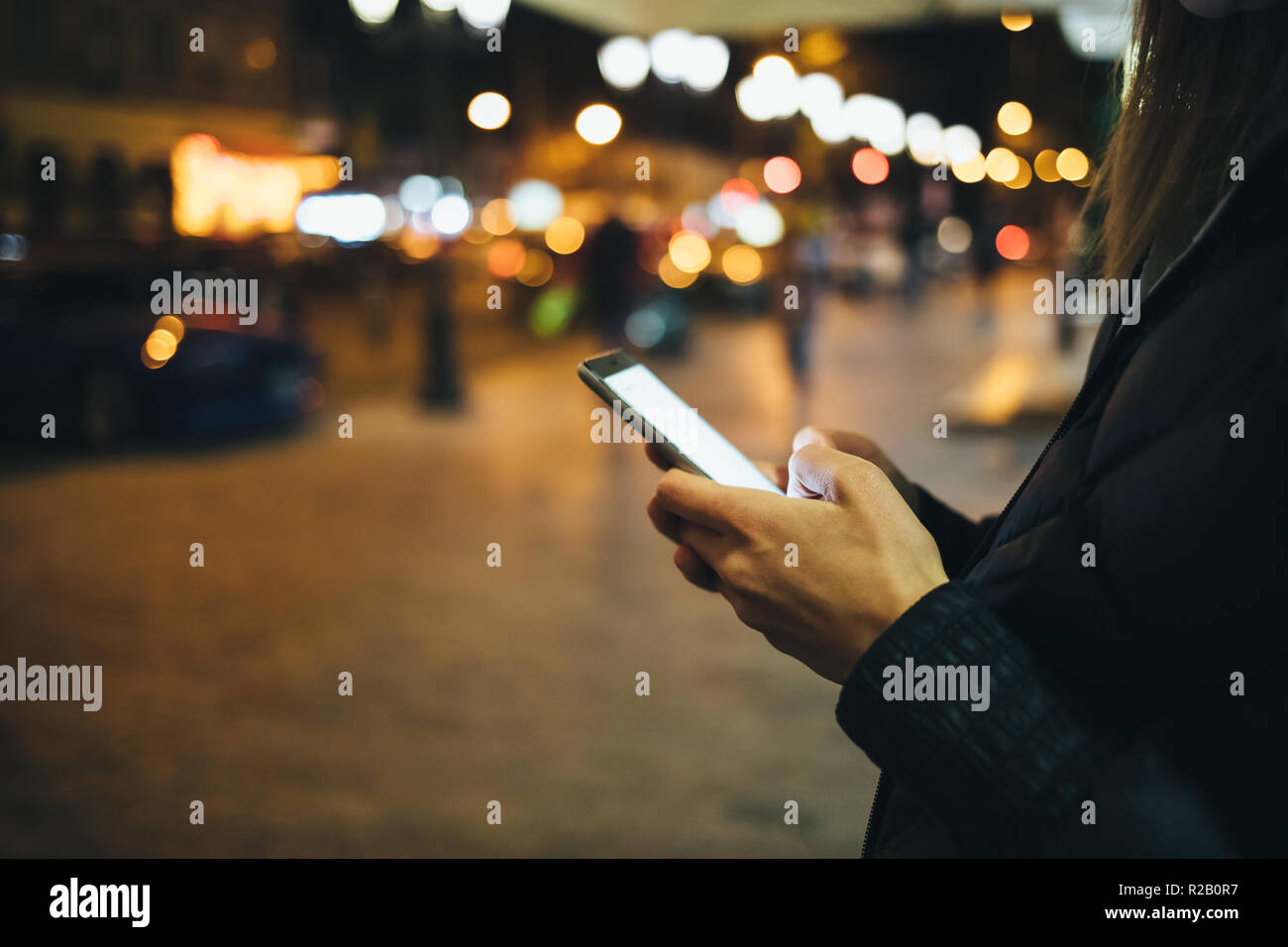 Close-up woman's hands using mobile phone with glowing screen on street, lights of night city on background. Stock Photo