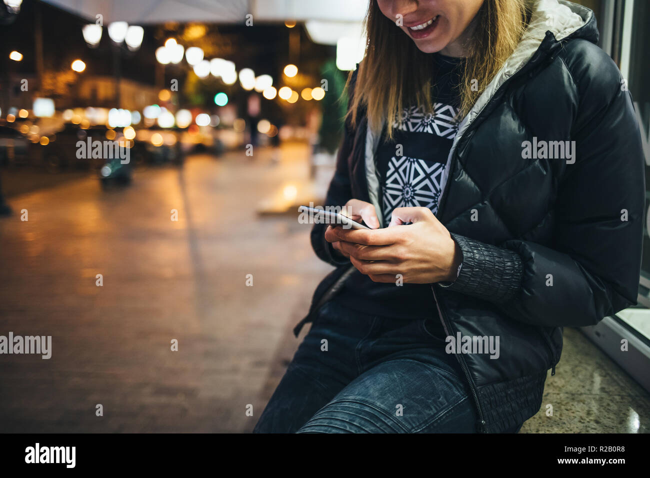 Happy young woman on street using mobile phone and smiles looking down at glowing screen, lights of night city on background. Stock Photo