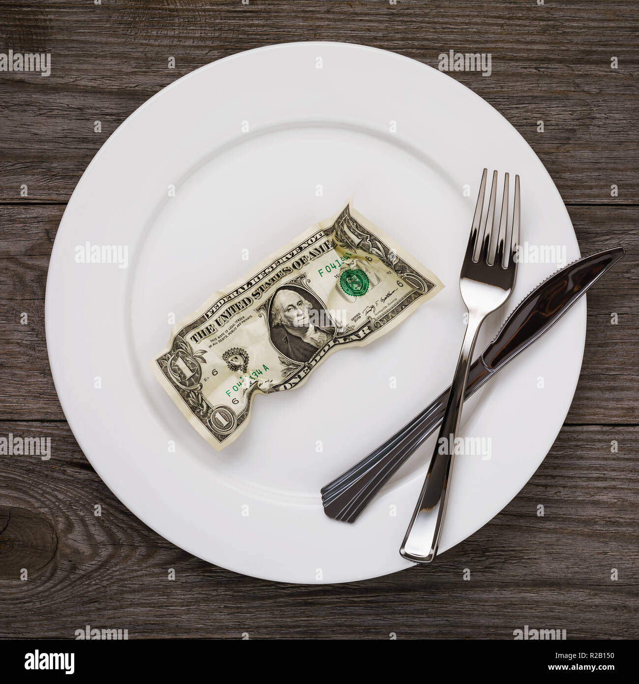 Crumpled dollar in the plate with fork and knife on the wooden table. Concept on the subject of human needs Stock Photo