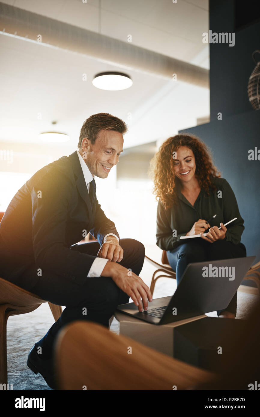 Two diverse young businesspeople smiling while stting in an office talking and working online together with a laptop Stock Photo